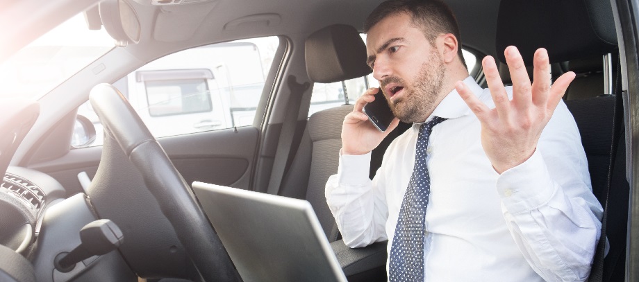 Stressed businessman seated in his car with laptop open, talking on a cell phone