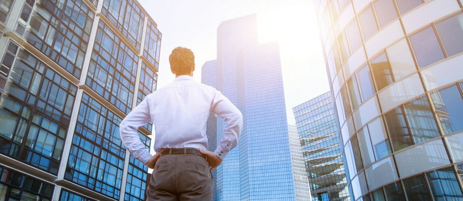 businessman viewed from behind as he contemplates a skyscraper with the sun behind it